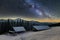 Old wooden house, hut and barn, pile of firewood in deep snow on mountain valley, spruce forest, woody hills on dark starry sky