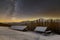 Old wooden house, hut and barn, pile of firewood in deep snow on mountain valley, spruce forest, woody hills on dark starry sky