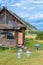 Old wooden house in mountains with solar panel on roof and milk cans on grass. The door is open. Wheelbarrow at the steps. Clouds