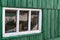 An old wooden small window with a frame of boards painted white and thin glass. On a battered wall of wooden planks with green pai