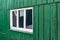 An old wooden small window with a frame of boards painted white and thin glass. On a battered wall of wooden planks with green pai