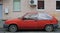 An old wrecked red hatchback car is parked outside the house