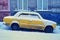 Old yellow car covered with snow parked on a snowy sloping street in winter city