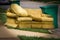 Old yellow couch with cushions sits on a curb in residential area next to a recycling cart waiting for bulk waste hauler pickup on