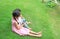 Older Asian sister hugs little brother by the neck, shoulders sitting on green grass field with playing smartphone together. Two