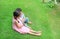 Older Asian sister hugs little brother by the neck, shoulders sitting on green grass field with playing smartphone together. Two