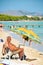 Older overweight man reading a newspaper at sandy beach under umbrella. People sunbathing at Playa de