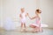 The older sister, a ballerina in a pink tutu and pointe shoes, shows the baby how to practice at the barre