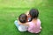 Older sister hugs little brother by the neck, shoulders sitting on green grass field with playing smartphone together. Two