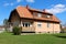 Older suburban family house with small balcony and wooden roof addition surrounded with grass and cloudy blue sky in background