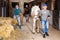 Older woman leading horse along stalls outdoor while young female worker stacking hay