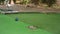 Older woman plays minigolf, detail only on her feet in gray sandals moving over green surface and blue ball moving to hole