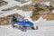 One blue snowmobile stands on snow close to stones in Norwegian mountains, warm day and bright sun, snow smelts, norwegian