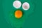 One broken white raw egg with yellow round yolk, two cracked halves of eggshell on green background isolated close up top view