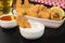 One fried tilapia strip in a bowl with tartar sauce close with a platter of fried tilapias and beer in black background