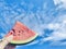 One hand holding a slice of watermelon, blue sky and white clouds background.