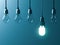 One hanging energy saving light bulb glowing different stand out from unlit incandescent bulbs with reflection on dark cyan backgr