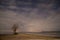 One lone tree on the lake shore at night with starry skies and clouds. Access road to the lake. One abandoned boat frozen in