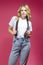 One Tranquil Winsome Caucasian Blond Girl In White Shirt Posing In Jeans While Standing Touching Suspenders Over Coral Pink