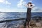 One Winsome Relaxing Caucasian Brunette Girl Posing in Black Dress With Lifted hands On Stone At Sea During Sunny Day Outdoors