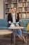 one woman sitting, 40 years old, holding magazine, book shop, book store, books on shelf behind out of focus