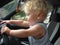 A one-year-old boy with magnificent blond hair in a summer T-shirt sits in the car in the driver`s seat and pretends that he is