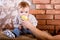 One year old child sits on a barrel against the background of a red brick wall and bit an green apple which is held by his father