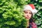 Ð¡onfused face woman in a santa claus red hat looks away through green fir tree branches in the forest. Christmas coming, sale