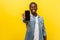 Online service, technology. Portrait of satisfied glad young man in denim casual shirt holding out cellphone. isolated on yellow