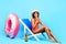 Oops. Shy black lady in bikini chilling in lounge chair, covering her mouth, making mistake on blue studio background
