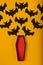 An open coffin with red padding from which black handmade paper bats fly out. halloween concept on yellow background