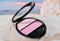 open face blush in pink shades and mini brush. decorative cosmetic on sandy beach next to shell. make up blusher for