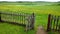 An open gate in a wooden fence and a green meadow beyond it, the path in the frame . Cloudy summer or late spring