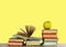 Open textbook, pile of books in colorful covers and green apple on wooden table with yellow background. Distance home education.