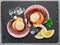 Opened fresh scallops on natural stone black slate plate with lemon slices. Flat lay