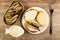 Opened jar with sprats, bowl with mayonnaise, slice of bread with mayonnaise, sandwich with sprats in plate, fork on wooden table