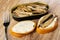 Opened jar with sprats, fork, slice of bread with mayonnaise, sandwich with sprats on wooden table