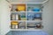 Opened white kitchen cupboard with plates, metal pots and food containers