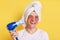Optimistic happy man wrapped towel on head doing morning cosmetology procedures, drying hair with hair dryer, laughing happily,