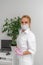 Optimistic portrait of a woman wearing a white coat. The blonde is standing next to a computer table wearing a mask and gloves.