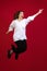 Optimistic young beautiful girl wearing white shirt laughing and jumping isolated on dark red studio background. Concept