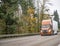 Orange big rig semi-truck transporting cargo in dry van semi trailer running on the foggy twilight highway road with autumn trees