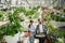 Orange colored watering device and box with plants. Florist man and woman are working together in bright greenhouse