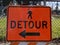 An orange detour sign on a walking path outside a chain link fence