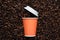 Orange disposable cup of coffee to go with a white lid on the background of roasted beans