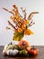 Orange Fall Flowers in a Vase with Little Orange, Green and White Gourds Lined Up In Front On a Wooden Table with a White Wall for