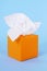 Orange kleenex style tissue box, white soft tissues, blue background, copy space