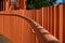 Orange railing with a shadow from the fence on a background of green trees and blue sky