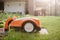 An orange robotic lawn mower stands on the base and is charged from electricity in the courtyard