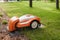 An orange robotic lawn mower stands on the base and is charged from electricity in the courtyard